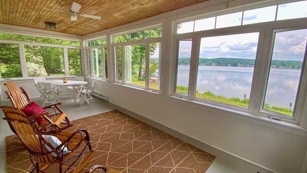 Un solárium con ventanas amplias que dan a un lago, amueblado con sillas y una mesa de madera.
