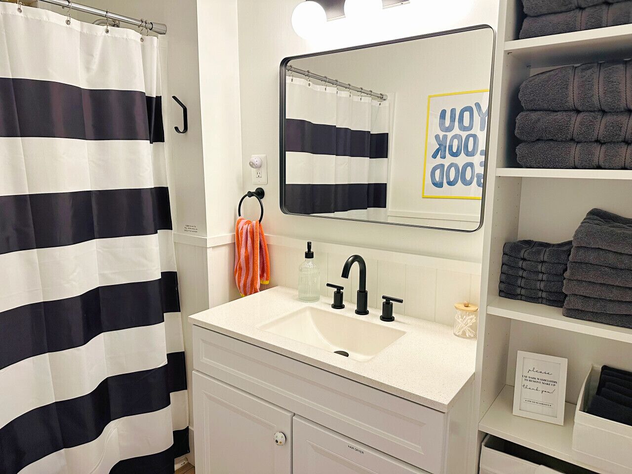 A bathroom with a white sink, black faucet, and a mirror above it. There is a striped shower curtain, a towel rack with a towel, and a shelf with folded towels.