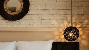 Une chambre avec un mur en briques, un miroir rond, un lit avec une literie blanche, un plaid noir et blanc et un luminaire suspendu.