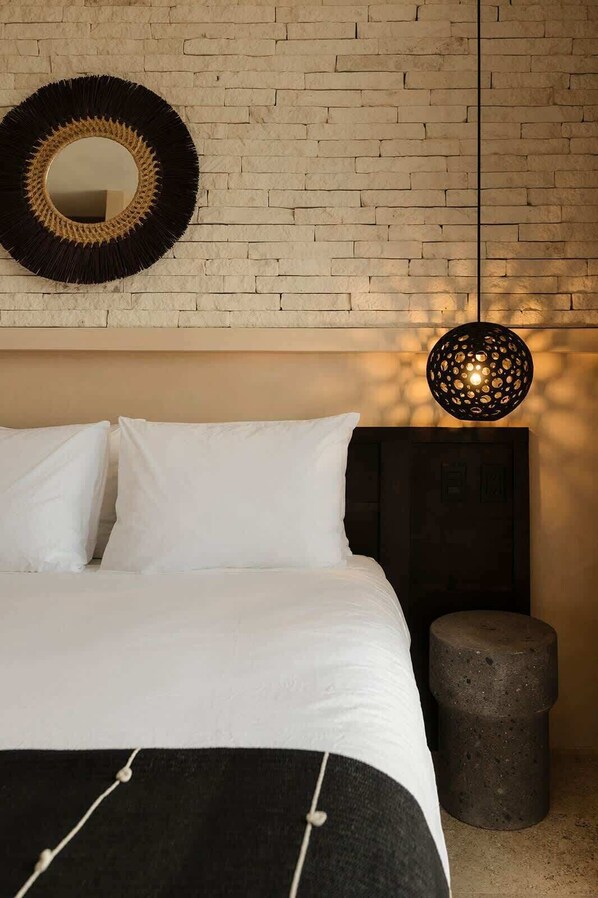 A bedroom with a brick wall, a round mirror, a bed with white bedding, a black and white throw, and a hanging light fixture.