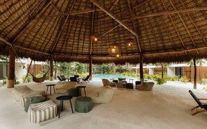 An open-air lounge with a thatched roof, outdoor seating, and a pool in the background. - K'Abax Jungle Apartment Beach Club Access (Tulum)