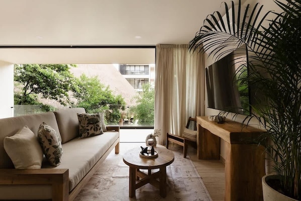 Una sala de estar moderna con un sofá, una mesa de centro y un televisor, que cuenta con un ventanal grande y una planta en maceta.