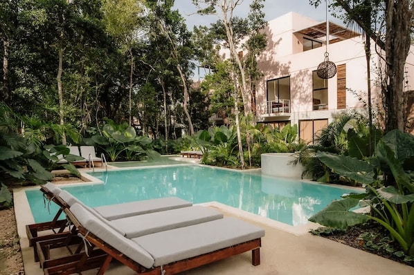 Ein Pool mit LiegestĂŒhlen, umgeben von ĂŒppiger Vegetation und einem modernen GebĂ€ude.