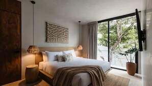 A modern bedroom with a large bed, a wall-mounted artwork, a potted plant, and a view of greenery outside.