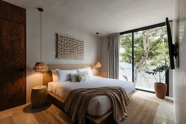 A modern bedroom with a large bed, a wall-mounted artwork, a potted plant, and a view of greenery outside.