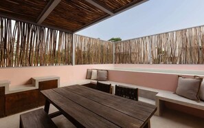 Een terras buiten met een houten tafel en bankjes, een vlechtwerk privacy scherm en een roze muur.