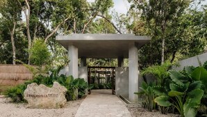 A stone pathway leading to a modern pavilion with a metal gate, surrounded by lush greenery and a large rock with 'SERMONIA HABITAT' inscribed.