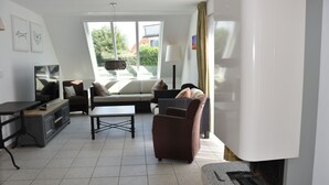 A modern living room with a sofa, armchair, and a dining table. There is a TV on a stand and a window with curtains.