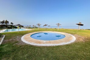 A circular pool with a surrounding grassy area, a view of the sea, and several thatched umbrellas. - Semi-detached house in second line beach in Manilva (Manilva)