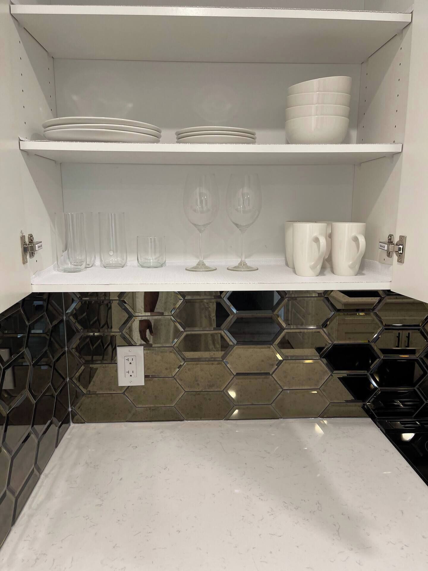 A kitchen with white shelves holding plates, glasses, and cups. Below the shelves, there's a countertop with a hexagonal tile backsplash.