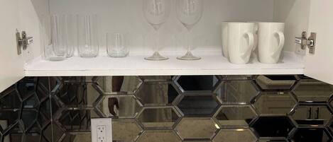 A kitchen with white shelves holding plates, glasses, and cups. Below the shelves, there's a countertop with a hexagonal tile backsplash.
