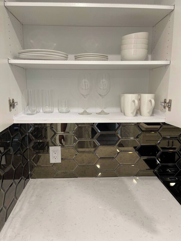 A kitchen with white shelves holding plates, glasses, and cups. Below the shelves, there's a countertop with a hexagonal tile backsplash. - Tastefully furnished one bedroom suite (Calgary)