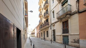 Una calle estrecha bordeada de edificios de varios pisos, con balcones de barandilla metálica, un camino peatonal despejado y una puerta visible a la izquierda.
