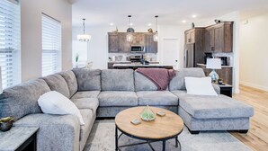 A modern living room with a sectional sofa, a round coffee table, and a kitchen in the background. - Comforable 3 bed 3 bath home (Cedar City)