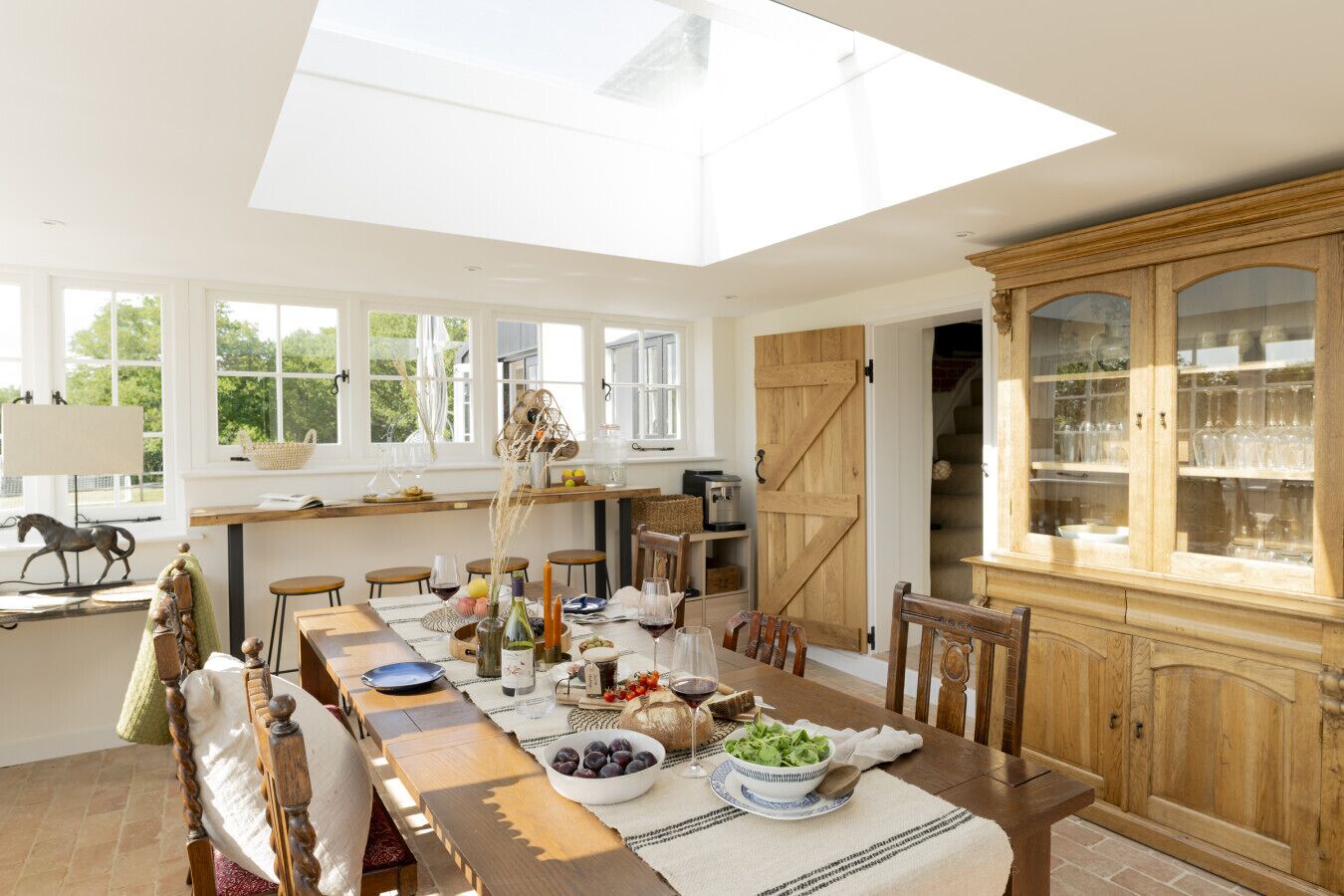 Un comedor con una larga mesa de madera, sillas, una araña y un mueble de madera grande con puertas de cristal.