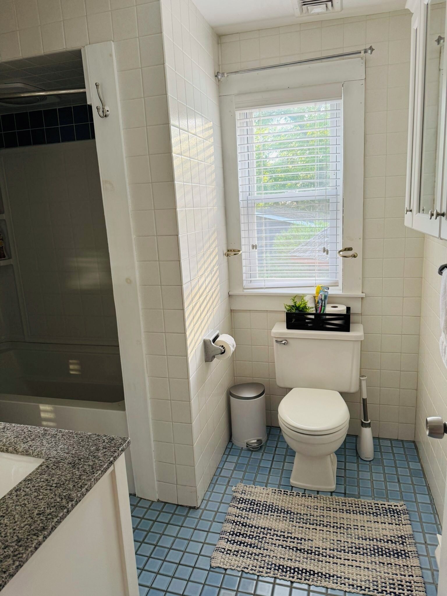 Ein Badezimmer mit Toilette, einem kleinen Mülleimer, einem Fenster mit Jalousien und einem Handtuchhalter.