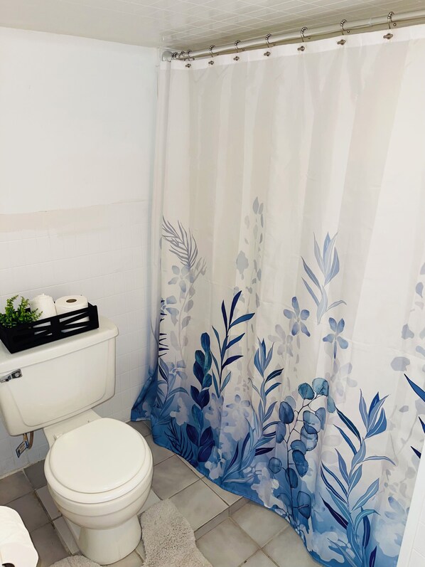 Une salle de bain avec des toilettes, un rideau de douche orné d’un motif floral bleu et un petit panier noir contenant des articles de toilette, posé sur le réservoir des toilettes.