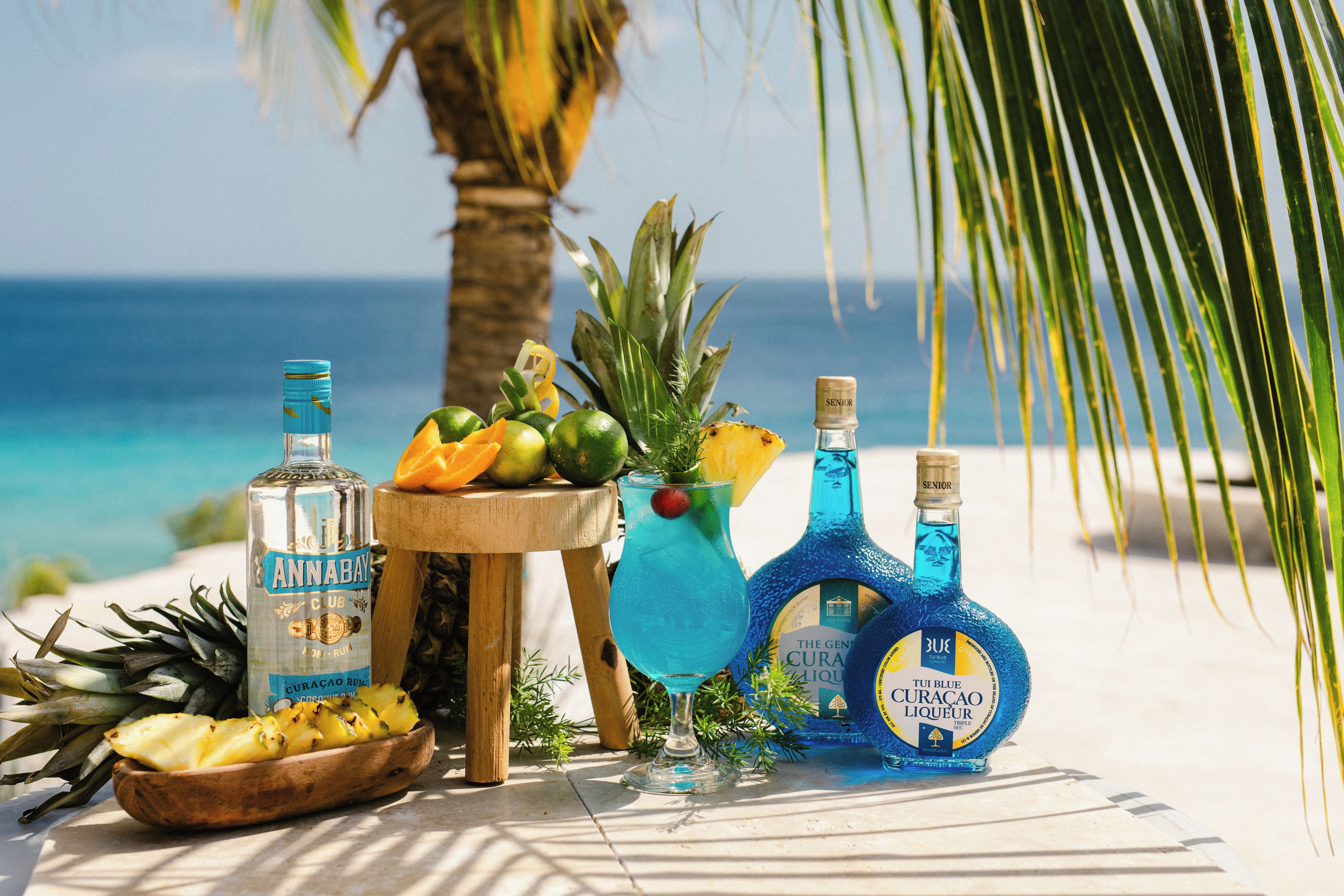 Un entorno tropical con una mesa de madera mostrando botellas de ron, un bol de rodajas de piña y una bebida tropical.