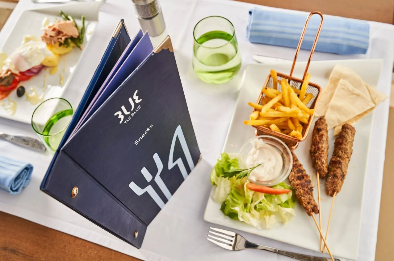 Pöydällä on lautanen, jossa on kebabia, ranskalaisia ja salaattia, lasi vihreää juomaa ja muistikirja, jonka kannessa lukee ”3L BLUE”.