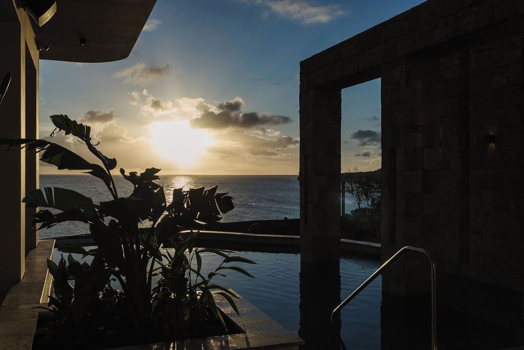 Vista do pôr do sol a partir de uma varanda com piscina e plantas tropicais.