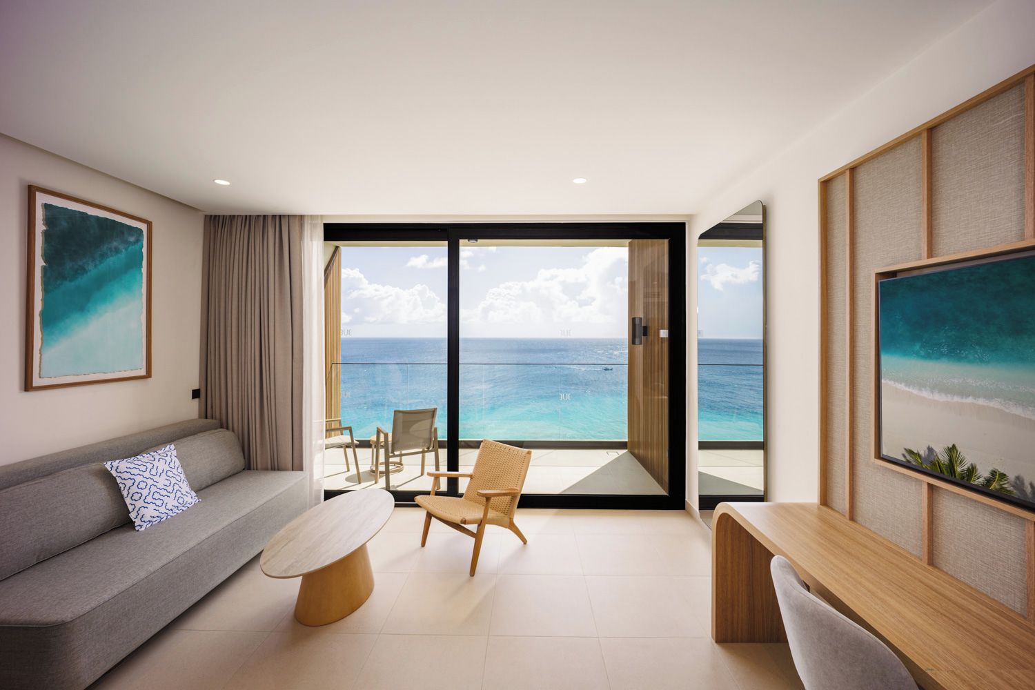 A modern living room with a sofa, a coffee table, and a chair, a balcony with a view of the ocean, and two framed artworks on the walls.