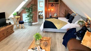 A loft-style bedroom with a sloped ceiling, a bed with multiple pillows, a wooden dresser, a small table with a plant, and a TV on a stand.