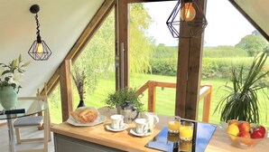 A kitchen with a view of a green landscape, a coffee maker, a kettle, a fruit bowl, and a glass of orange juice.