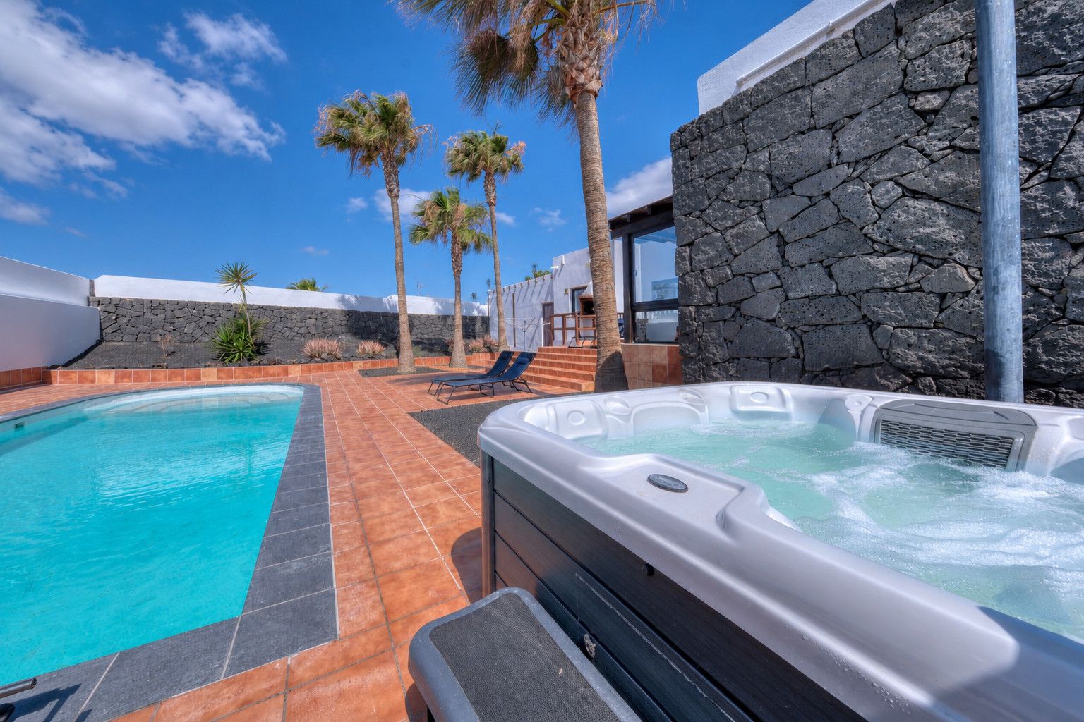 A swimming pool with a hot tub, surrounded by palm trees and stone walls.