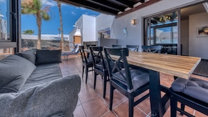 An outdoor patio with a wooden table and chairs, a gray sofa, and a view of the beach.