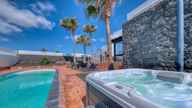 A swimming pool, a hot tub, and a stone wall with palm trees in the background.