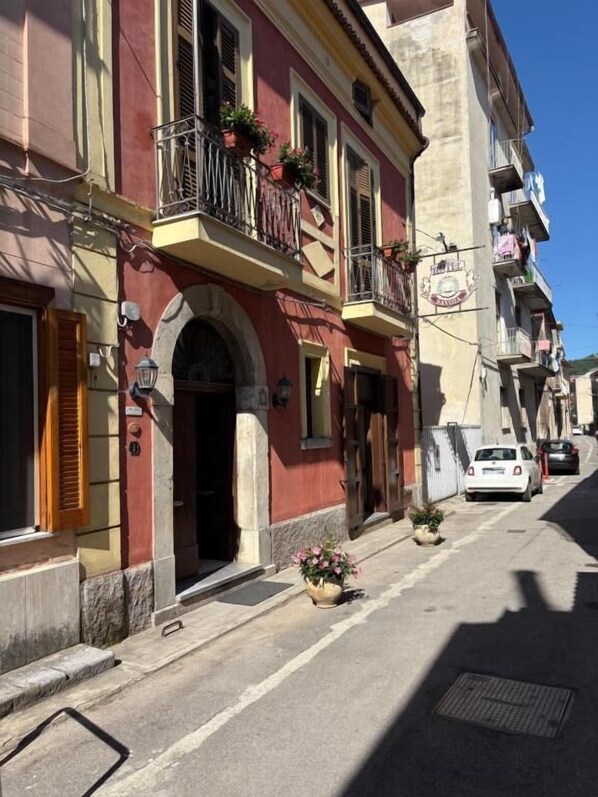 Una calle estrecha con edificios que tienen balcones con plantas en macetas, un cielo azul despejado y un coche blanco estacionado.