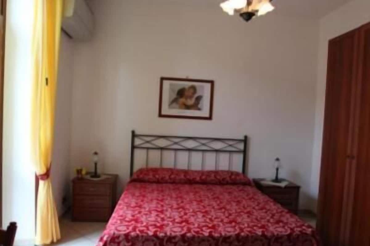 Un dormitorio con cama, mesita de noche, un cuadro en la pared y ventana con cortinas.