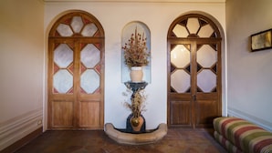 Un vestíbulo con tres puertas de madera y vidrieras, un jarrón central con flores secas, y un banco a la derecha.