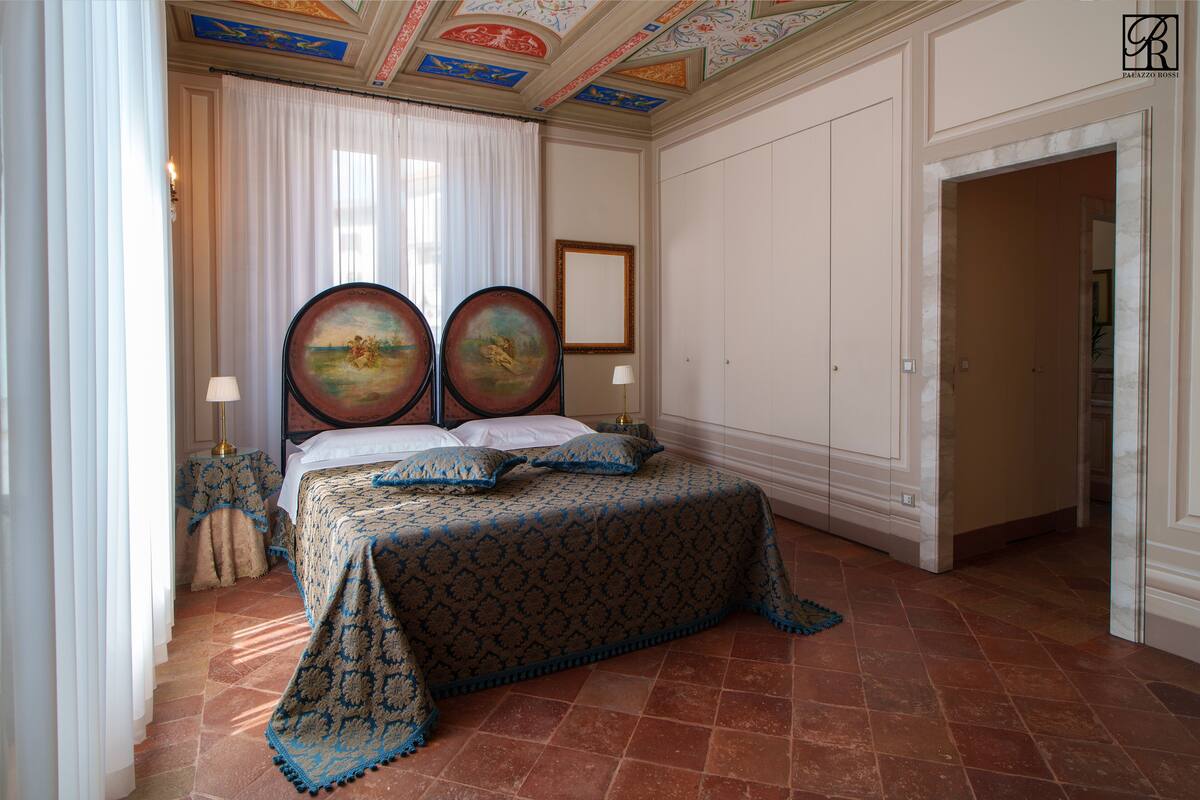 A bedroom with a large bed, two wall-mounted paintings, a white curtain, and a tiled floor.
