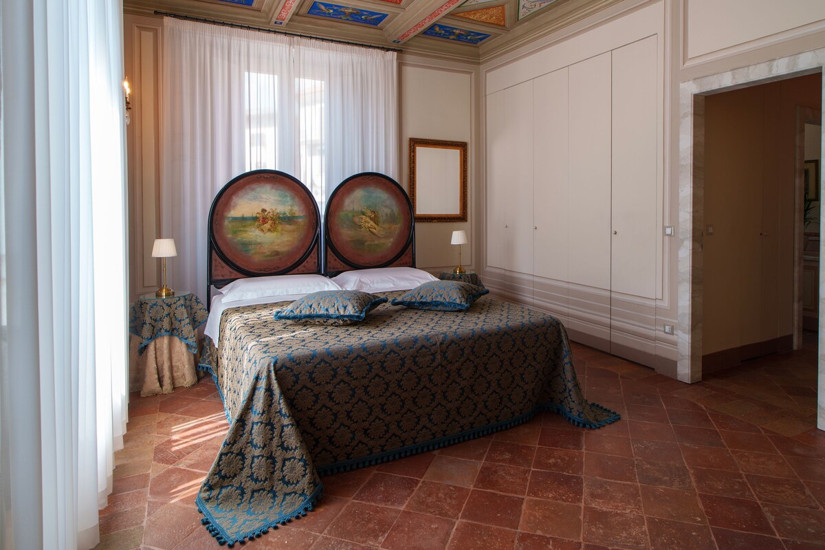 A bedroom with a large bed, two wall-mounted paintings, a white curtain, and a tiled floor.