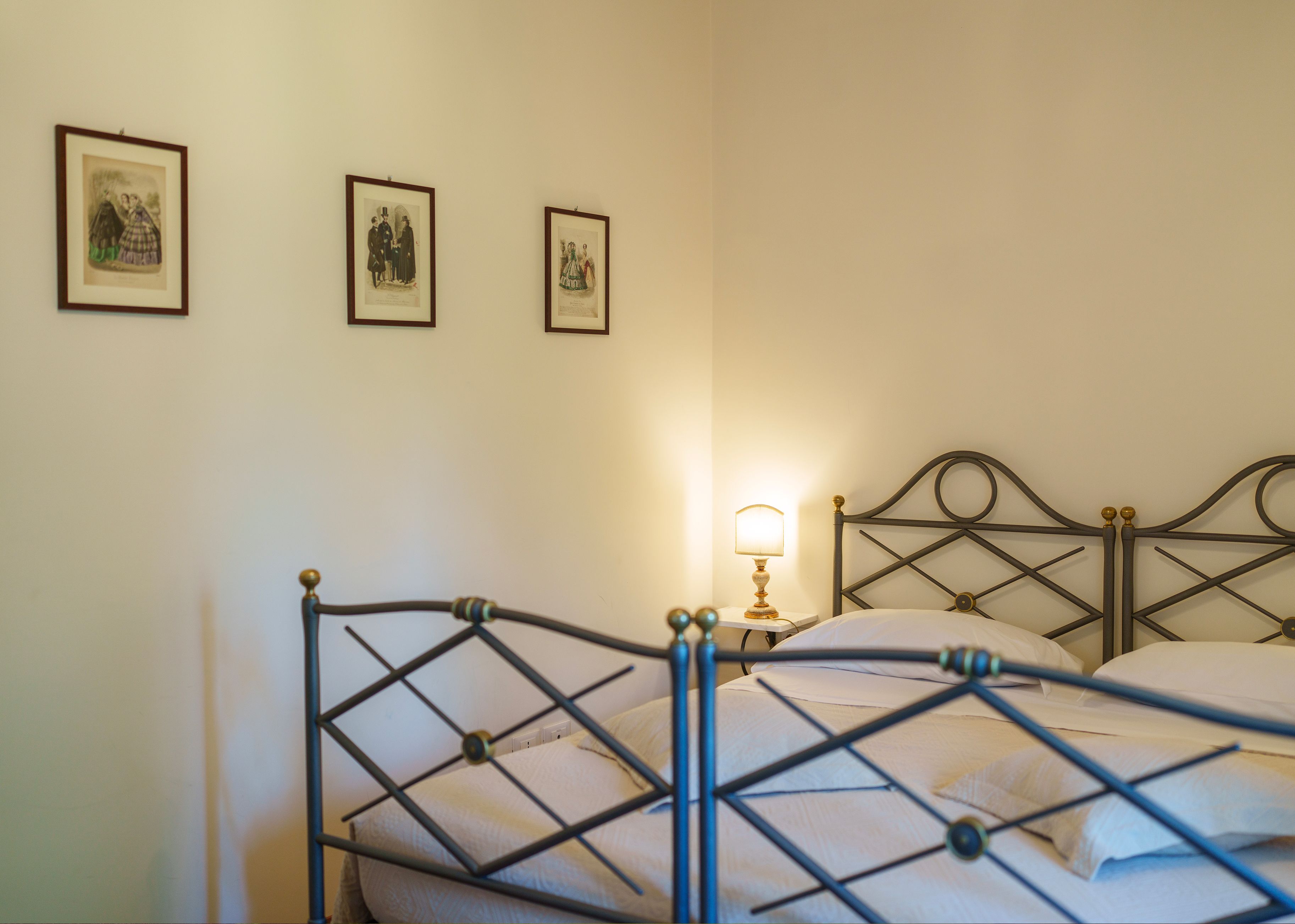 A neatly made bed with a metal frame, a bedside table with a lamp, and three framed pictures on the wall.