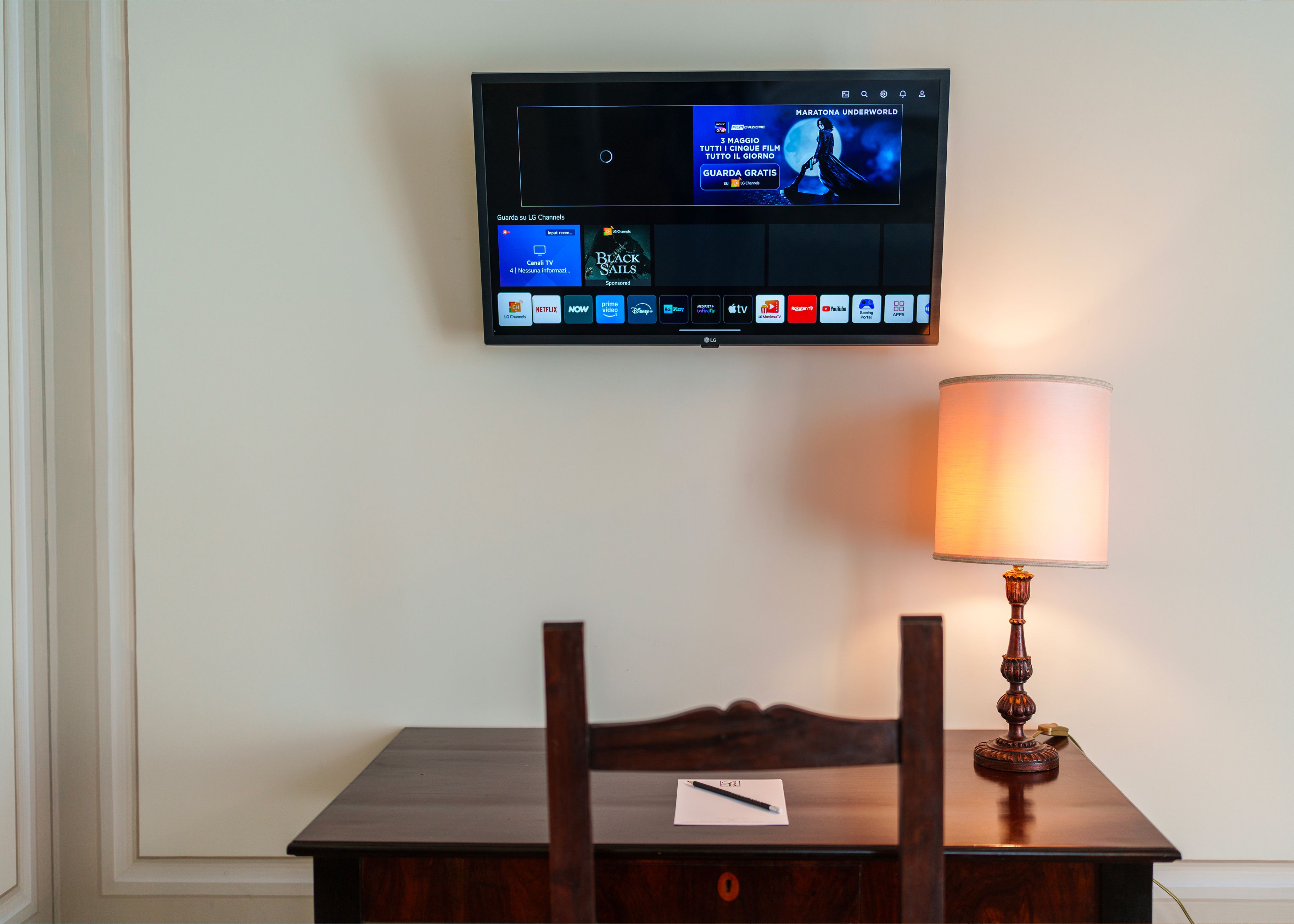 Een flatscreen-tv aan de muur waarop een film draait, een lamp op een tafel en een stoel met een notitieboekje.