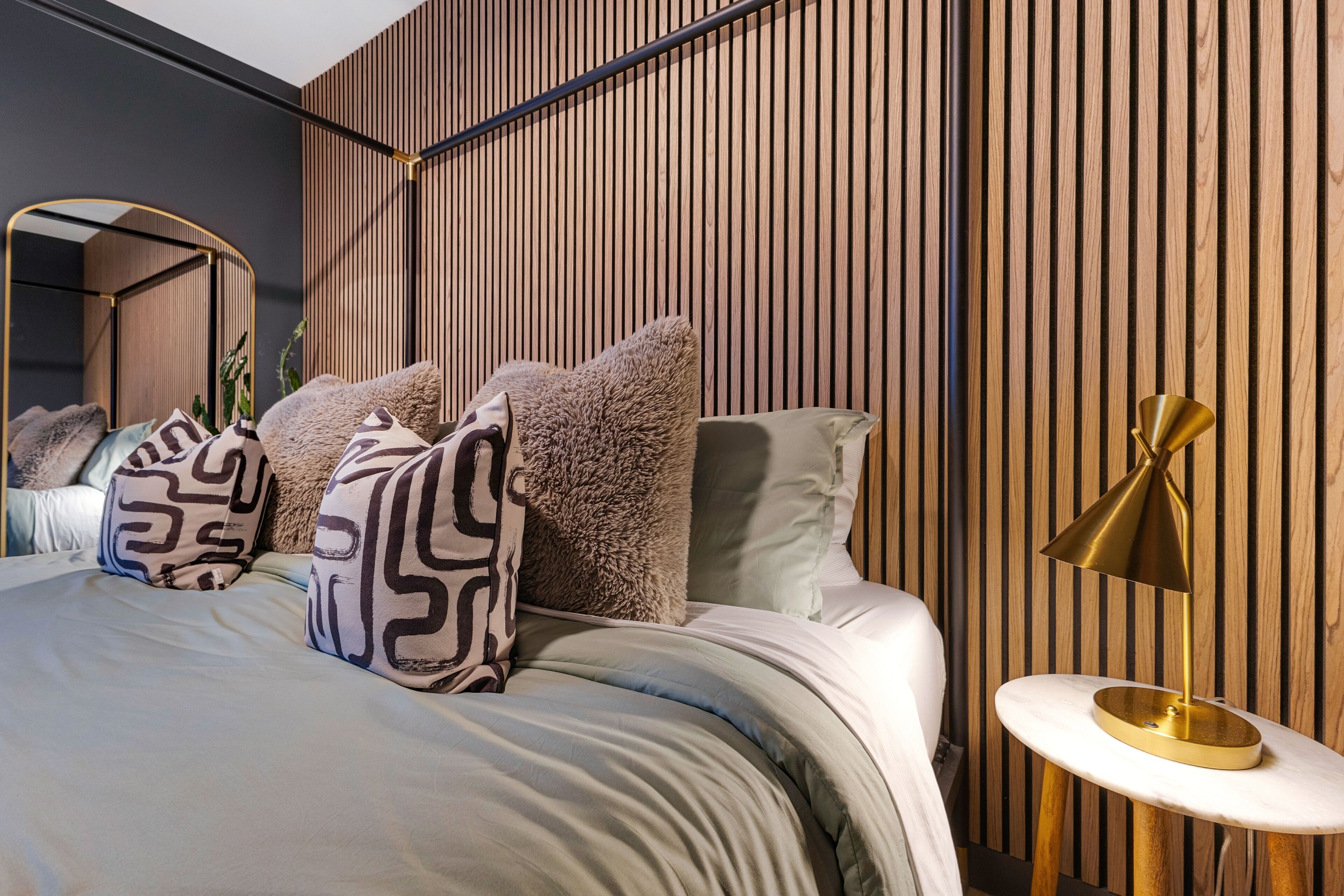 A modern bedroom with a bed, pillows, a bedside table with a lamp, and a wooden slatted wall.