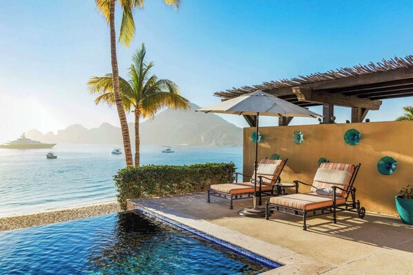 A poolside area with lounge chairs, a pergola, and a view of the sea with boats.