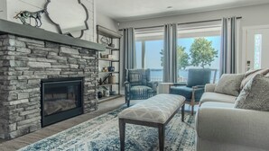A living room with a stone fireplace, a blue patterned rug, a blue armchair, a beige sofa, and a view of the outdoors. - Modern Lakeside Retreat on Lake Ontario 4+ bedroom/3 bath (Dexter)