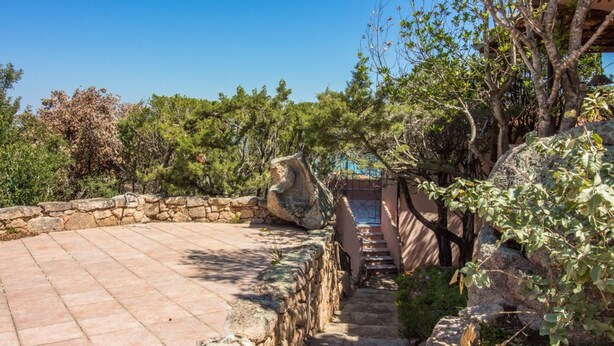 Un sendero de piedra que conduce a un patio circular rodeado de follaje y un muro de piedra.
