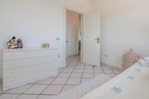 Een slaapkamer met een witte dressoir, een bed met een beddegoed met een patroon, een rieten mand en een betegelde vloer.