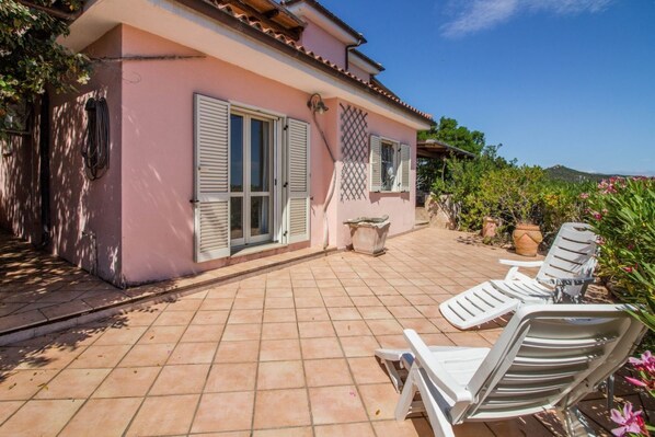Eine pinkfarbene Villa mit einer gefliesten Terrasse, zwei Fenstern mit Rollläden und einem kleinen Garten mit Pflanzen und einem Topfbaum.