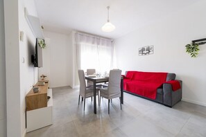 A modern living room with a dining area, a grey sofa with a red blanket, a TV mounted on the wall, and a window with sheer curtains.