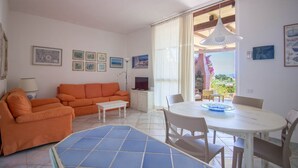 A living room with a sofa, a small table, and a dining table with chairs. There are framed pictures on the wall and a window with curtains. - GuestHost - Seaview Villa with Veranda GolfoAranci (Golfo Aranci)