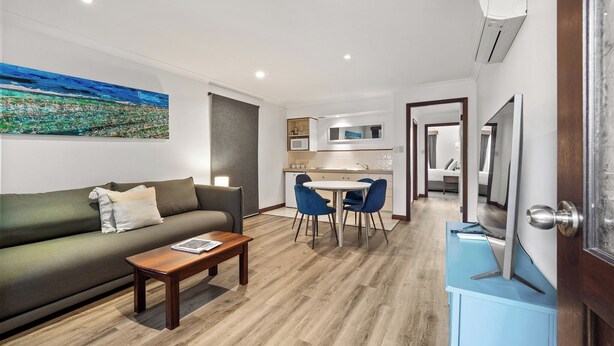 A modern living room with a sofa, coffee table, and a dining area with chairs. There is a kitchen in the background with a microwave and cabinets.