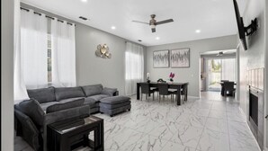 A living room with a sectional sofa, a dining table with chairs, a ceiling fan, and a fireplace.