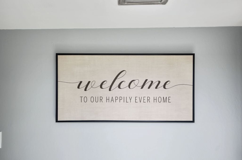 A framed welcome sign with the text "welcome to our happily ever home" on a light gray wall.