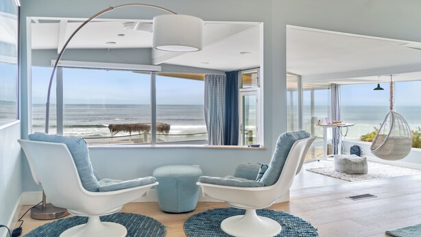 Una sala de estar moderna con ventanas amplias que dan al océano, dos sillas blancas con cojines azules, una silla colgante y una mesita con una lámpara.