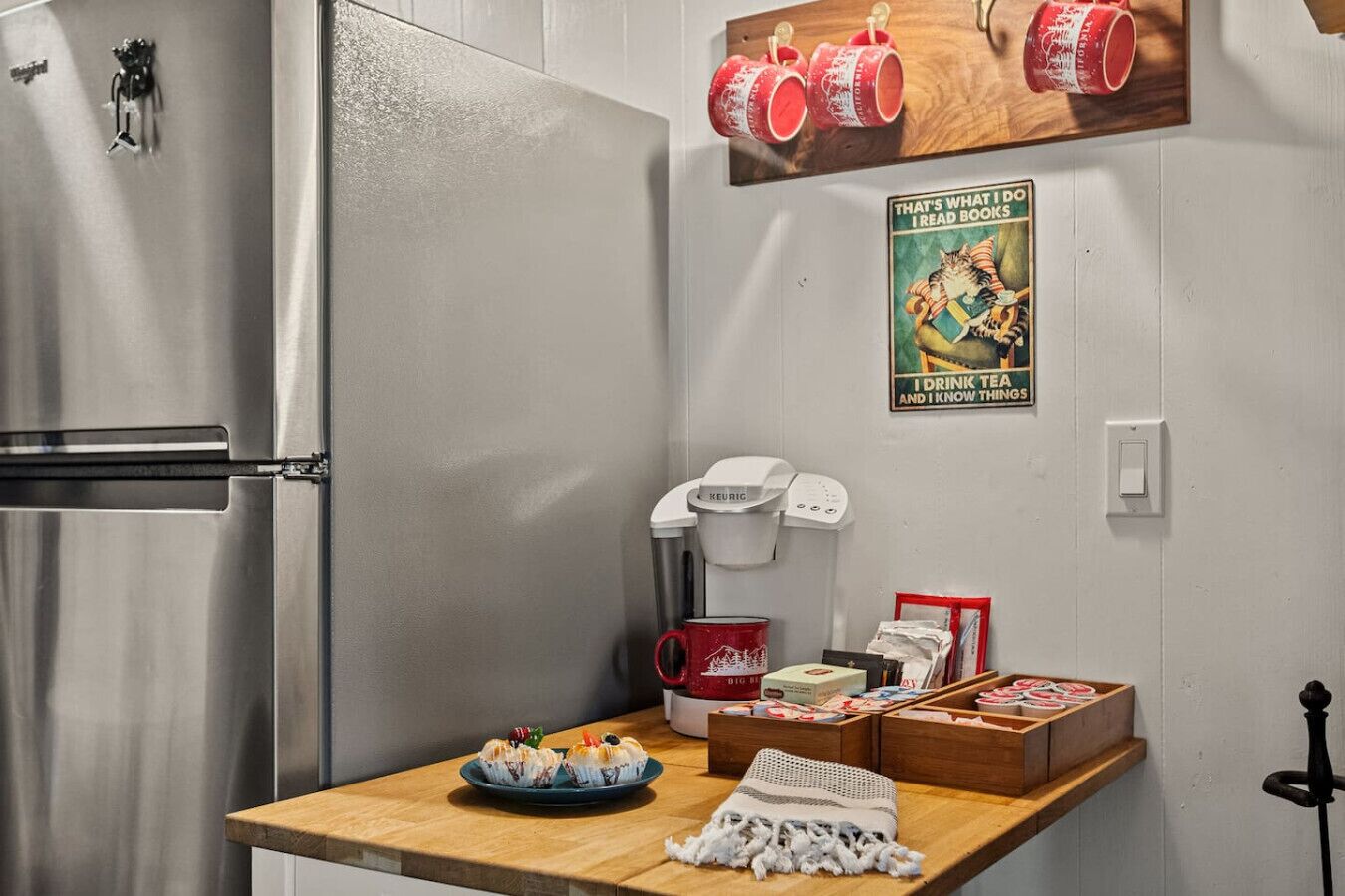 A kitchen with a stainless steel refrigerator, a coffee maker, and a wooden shelf with mugs and books.